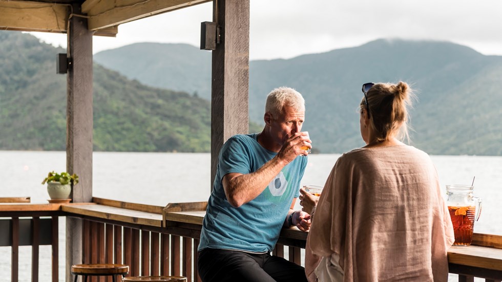Couple relax at the Boatshed Cafe and Bar outdoor jetty bar with drinks and views of the scenic Endeavour Inlet from Punga Cove