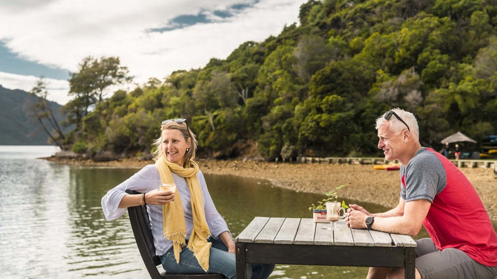 A relaxed couple drink and enjoy Boatshed Cafe and Bar coffee on the jetty at Punga Cove in the Marlborough Sounds in New Zealand