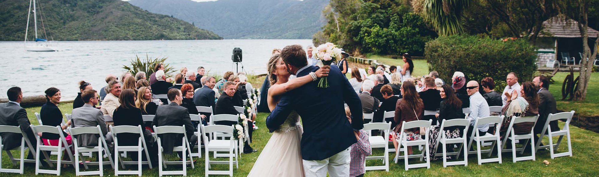 A newly married bride and groom kiss on the front lawn of Punga Cove following the ceremony with watching guests in the Marlborough Sounds of New Zealand