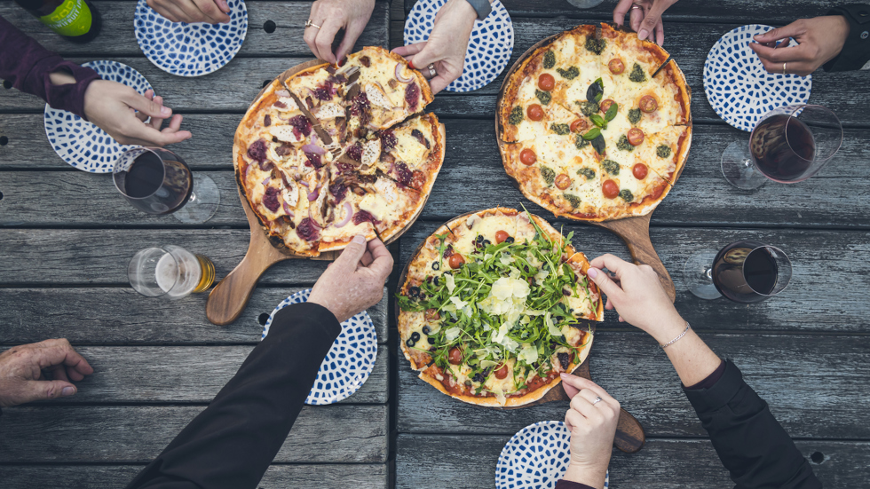 A group of friends share three hot stone baked pizzas and drinks from the Boatshed Cafe and Bar at Punga Cove located in the Marlborough Sounds of New Zealand's South Island