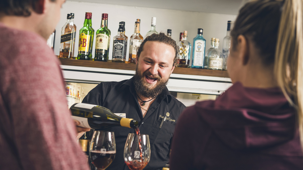 A friendly bartender pours Pinot Noir into glasses for customers at the Boatshed Cafe and Bar at Punga Cove - choose from a great range of local craft beer, wine , cocktails, soft drinks and expresso coffee.